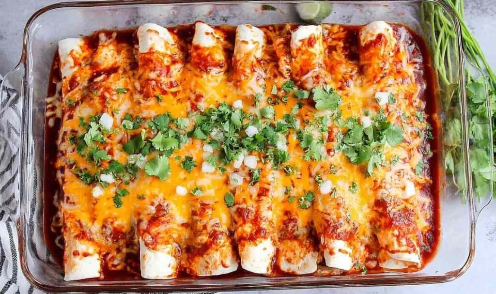 A pan of baked chicken enchiladas covered in melted cheese and red enchilada sauce, garnished with fresh cilantro and a dollop of sour cream