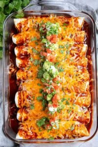 A pan of baked chicken enchiladas covered in melted cheese and red enchilada sauce, garnished with fresh cilantro and a dollop of sour cream