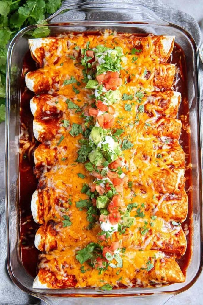A pan of baked chicken enchiladas covered in melted cheese and red enchilada sauce, garnished with fresh cilantro and a dollop of sour cream