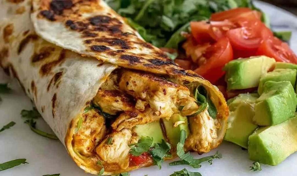 A sliced chicken avocado wrap on a white plate showing layers of tender chicken, fresh avocado, lettuce, tomato, and cheddar cheese inside a flour tortilla
