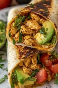 A sliced chicken avocado wrap on a white plate showing layers of tender chicken, fresh avocado, lettuce, tomato, and cheddar cheese inside a flour tortilla