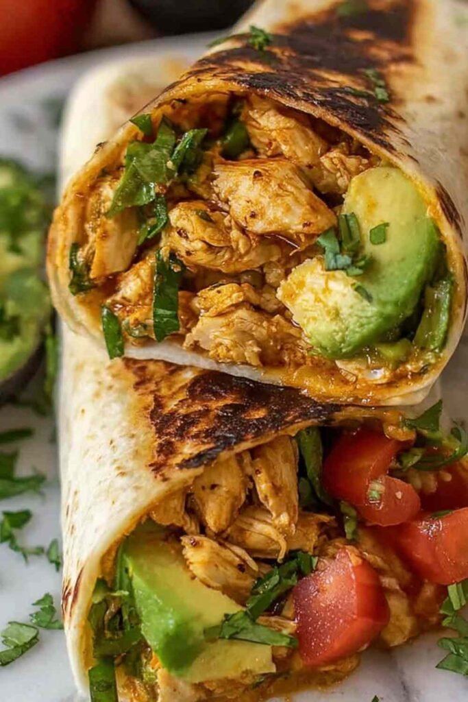 A sliced chicken avocado wrap on a white plate showing layers of tender chicken, fresh avocado, lettuce, tomato, and cheddar cheese inside a flour tortilla