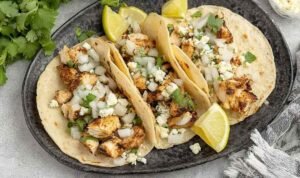 Close-up of assembled chicken street tacos on a wooden board with white onion, fresh cilantro, and lime wedges