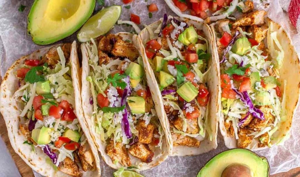 Overhead view of chicken tacos filled with seasoned chopped chicken, shredded lettuce, tomato, red onion, and avocado in flour tortillas