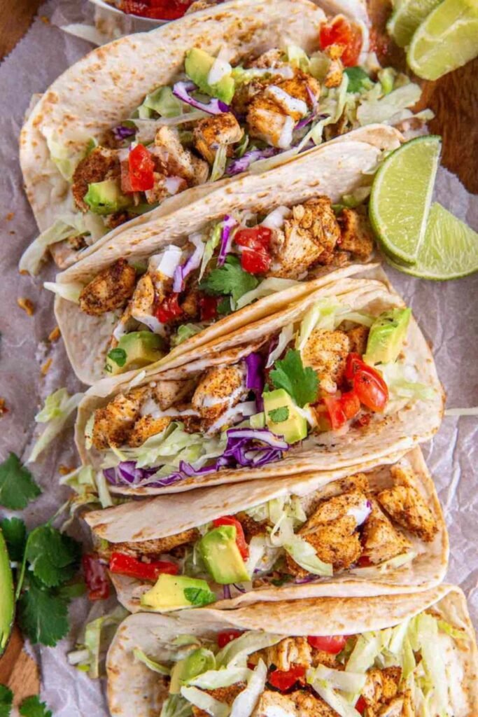 Overhead view of chicken tacos filled with seasoned chopped chicken, shredded lettuce, tomato, red onion, and avocado in flour tortillas