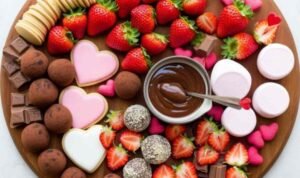 Overhead view of a Chocolate and Strawberry Dessert Board on a round wooden board with fresh strawberries, truffles, broken chocolate bars, heart cookies, pink marshmallows, and a chocolate dipping sauce bowl in the center