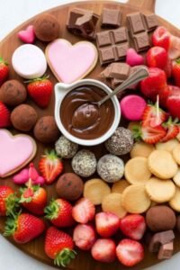 Overhead view of a Chocolate and Strawberry Dessert Board on a round wooden board with fresh strawberries, truffles, broken chocolate bars, heart cookies, pink marshmallows, and a chocolate dipping sauce bowl in the center