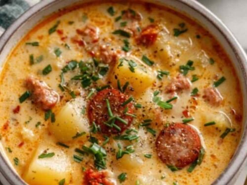 Creamy crockpot potato soup with sausage in a white bowl topped with cheddar cheese, crumbled bacon, and green onions