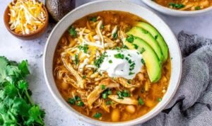 Crockpot White Chicken Chili A bowl of crockpot white chicken chili topped with avocado slices, chopped cilantro, and tortilla chip strips on a wooden table