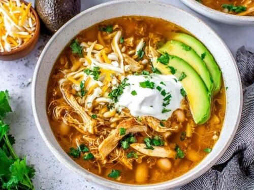 A bowl of crockpot white chicken chili topped with avocado slices, chopped cilantro, and tortilla chip strips on a wooden table