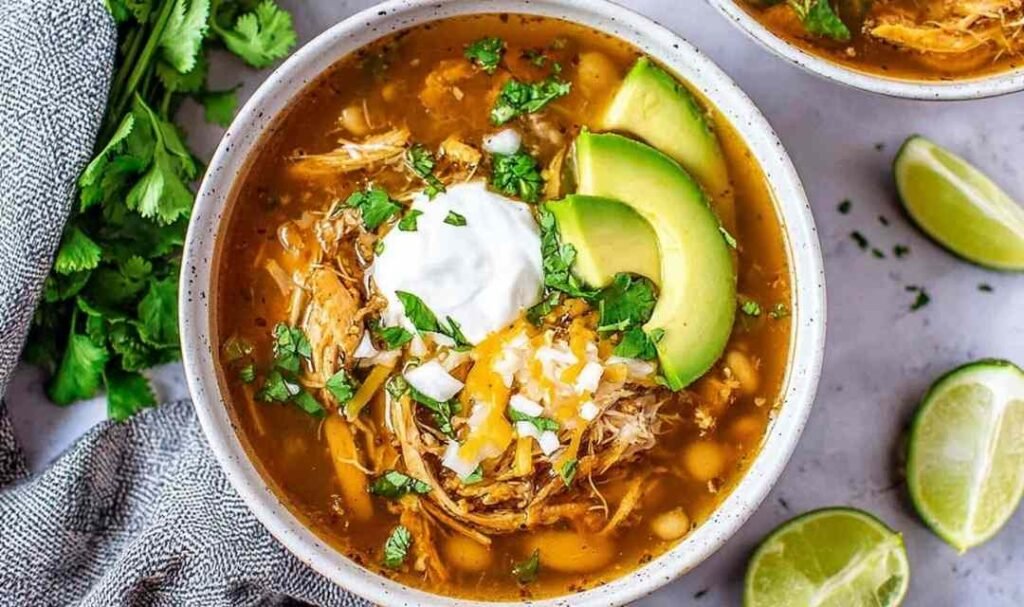 A bowl of crockpot white chicken chili topped with avocado slices, chopped cilantro, and tortilla chip strips on a wooden table