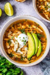 A bowl of crockpot white chicken chili topped with avocado slices, chopped cilantro, and tortilla chip strips on a wooden table