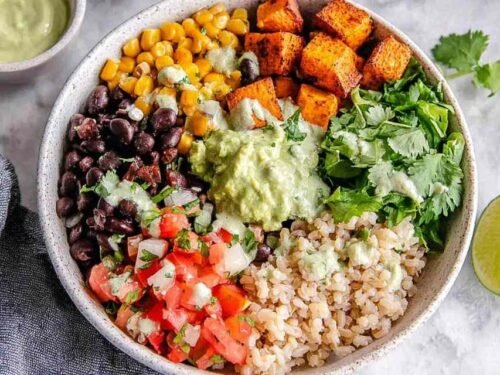 A colorful vegan burrito bowl with brown rice, roasted sweet potato, black beans, corn, and guacamole