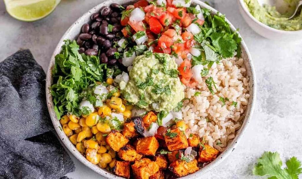 A colorful vegan burrito bowl with brown rice, roasted sweet potato, black beans, corn, and guacamole