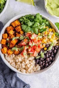 A colorful vegan burrito bowl with brown rice, roasted sweet potato, black beans, corn, and guacamole