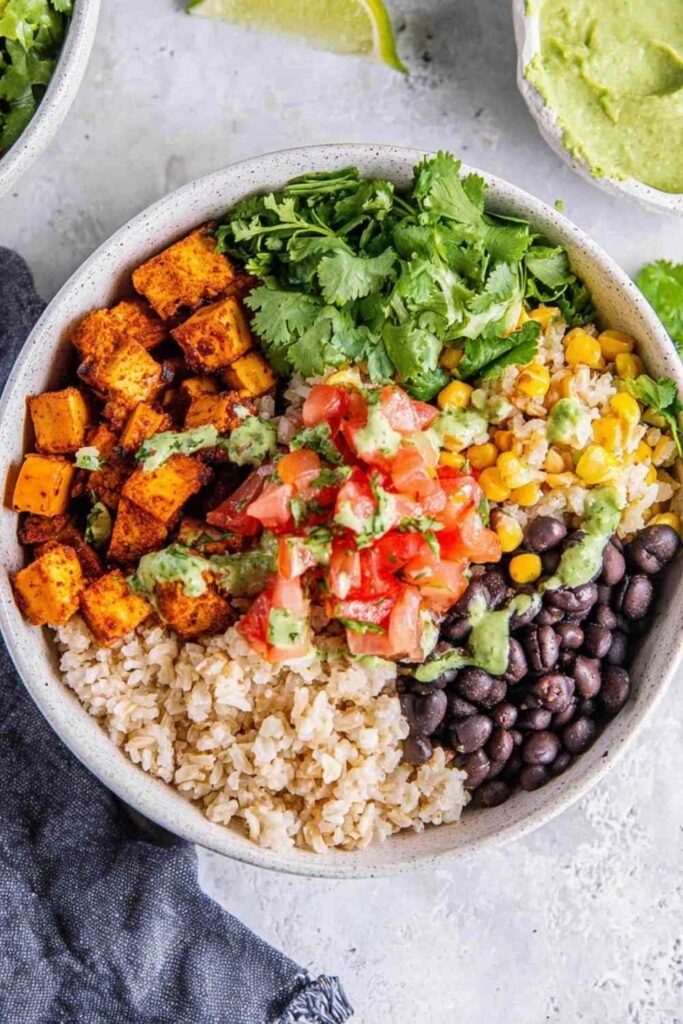A colorful vegan burrito bowl with brown rice, roasted sweet potato, black beans, corn, and guacamole