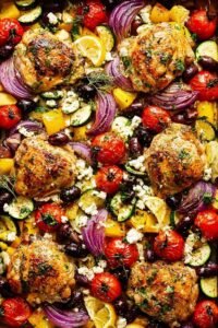 Greek sheet pan chicken with roasted vegetables, kalamata olives, and crumbled feta on a rimmed baking sheet