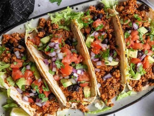 Four ground turkey tacos topped with lettuce, cilantro, and red onion on a white plate