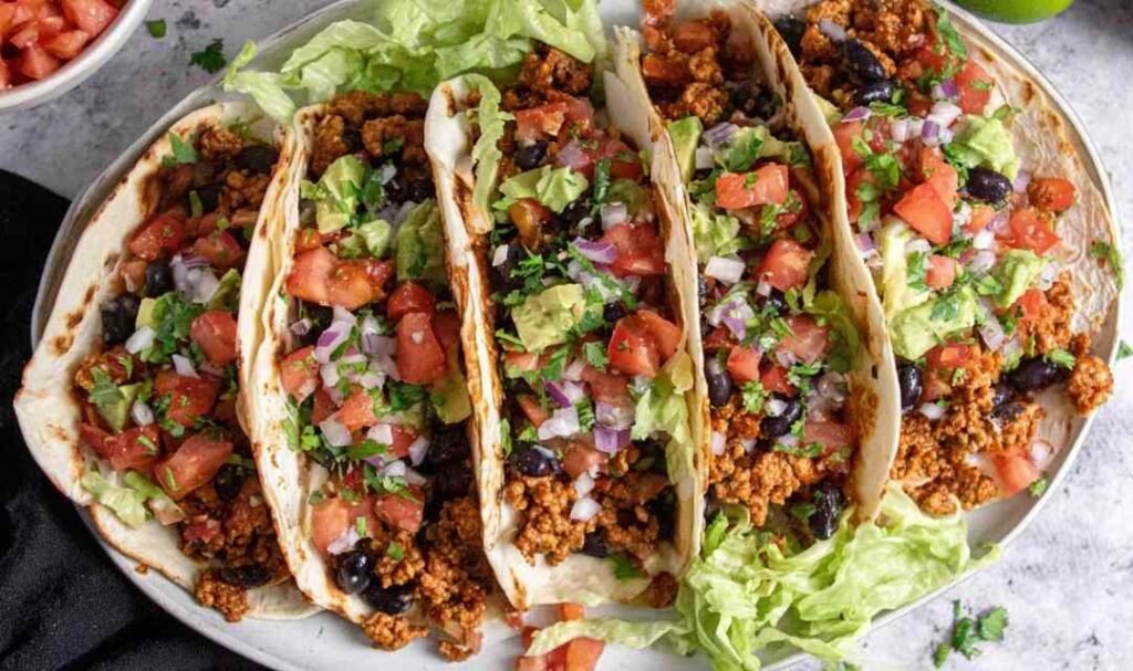 Four ground turkey tacos topped with lettuce, cilantro, and red onion on a white plate