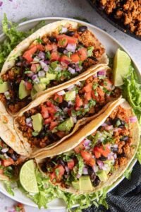 Four ground turkey tacos topped with lettuce, cilantro, and red onion on a white plate