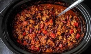 A hearty bowl of slow cooker chili topped with shredded cheddar cheese, sour cream, and sliced avocado