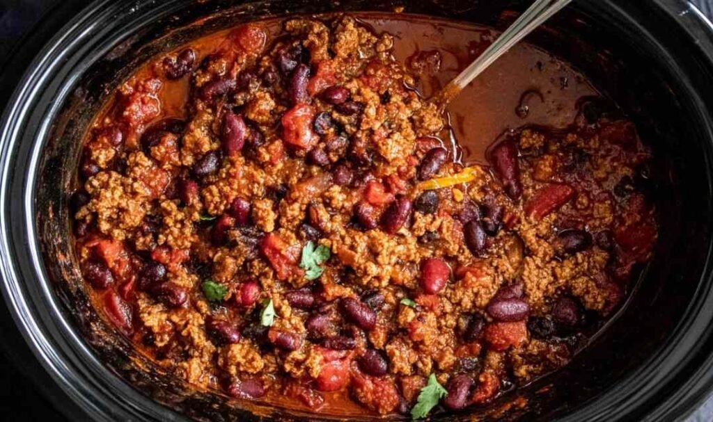 A hearty bowl of slow cooker chili topped with shredded cheddar cheese, sour cream, and sliced avocado