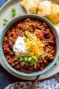 A hearty bowl of slow cooker chili topped with shredded cheddar cheese, sour cream, and sliced avocado
