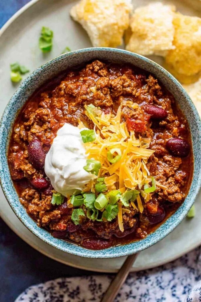 A hearty bowl of slow cooker chili topped with shredded cheddar cheese, sour cream, and sliced avocado