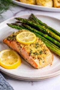Four lemon garlic baked salmon fillets in a parchment-lined baking dish topped with fresh herbs