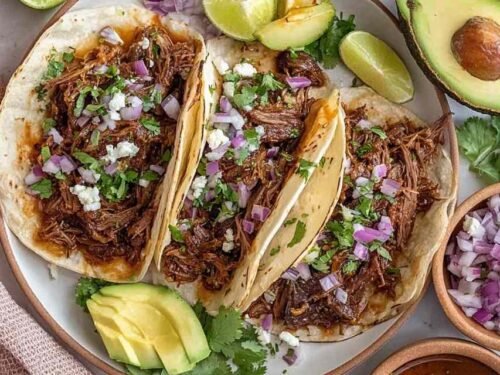 Mexican shredded beef tacos in warm corn tortillas topped with fresh cilantro, chopped white onion, and sliced avocado