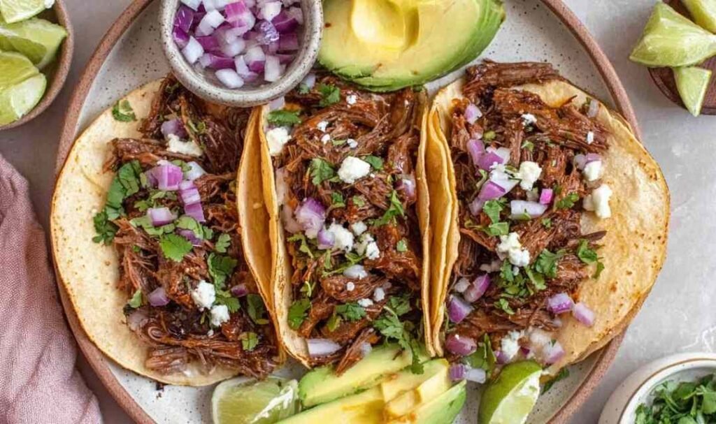 Mexican shredded beef tacos in warm corn tortillas topped with fresh cilantro, chopped white onion, and sliced avocado