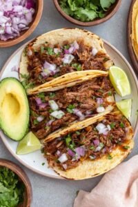 Mexican shredded beef tacos in warm corn tortillas topped with fresh cilantro, chopped white onion, and sliced avocado
