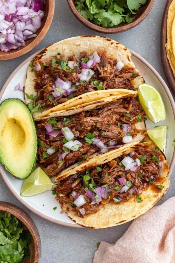 Mexican shredded beef tacos in warm corn tortillas topped with fresh cilantro, chopped white onion, and sliced avocado