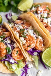 Shrimp Tacos on a plate with crunchy red cabbage, avocado slices, Cotija cheese, and creamy spicy sauce drizzle