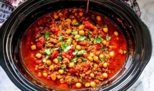 Slow Cooker Turkey Chickpea Chili A bowl of slow cooker turkey chickpea chili topped with sliced green onions, avocado, and crumbled tortilla chips on a wooden surface