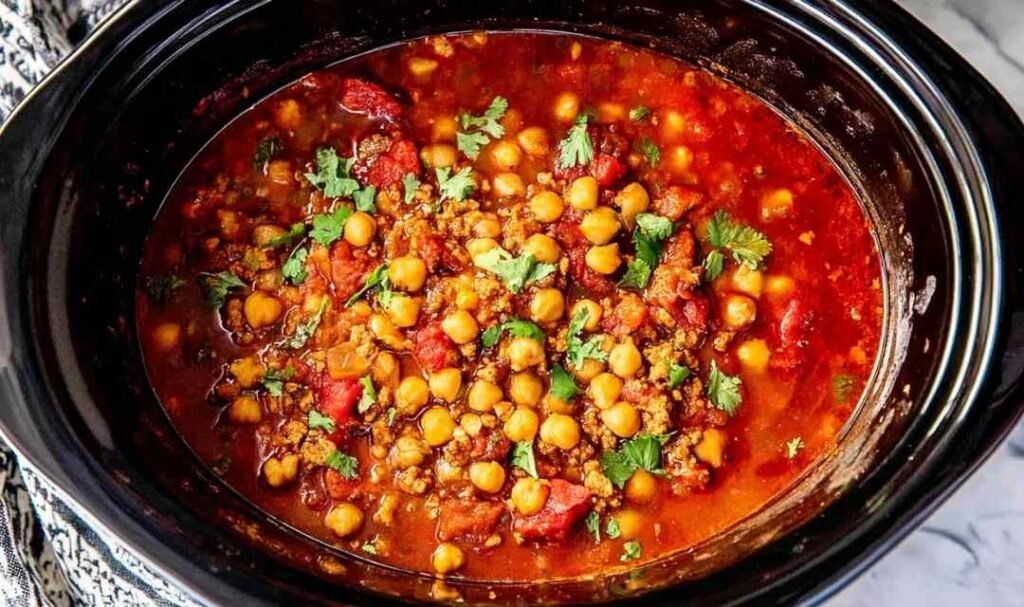 A bowl of slow cooker turkey chickpea chili topped with sliced green onions, avocado, and crumbled tortilla chips on a wooden surface