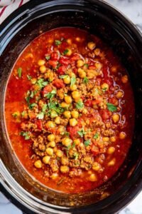 A bowl of slow cooker turkey chickpea chili topped with sliced green onions, avocado, and crumbled tortilla chips on a wooden surface