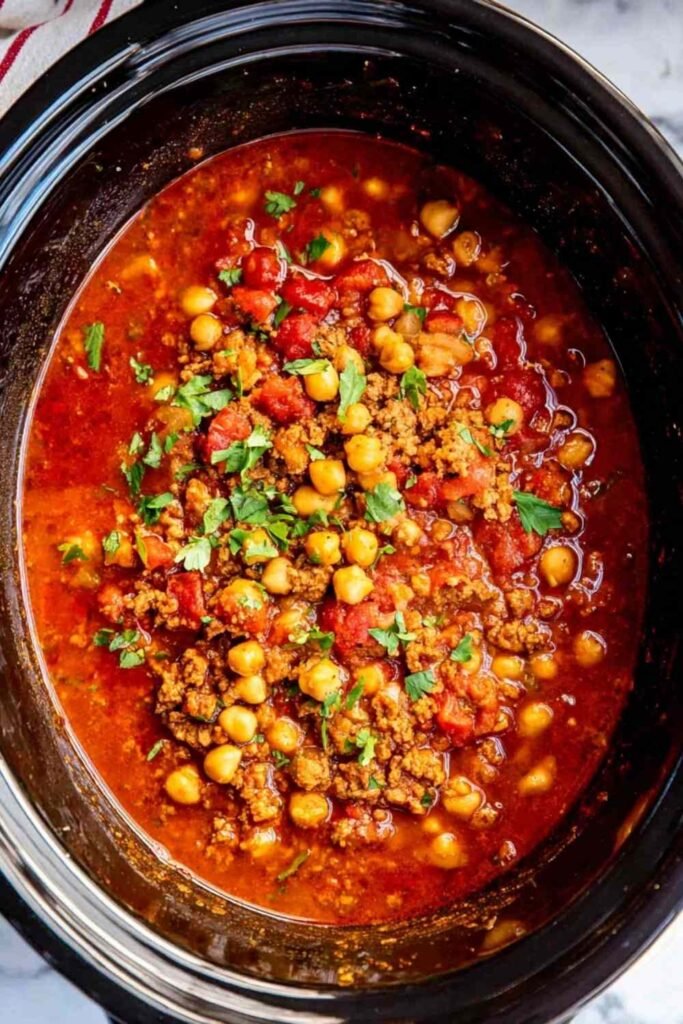 A bowl of slow cooker turkey chickpea chili topped with sliced green onions, avocado, and crumbled tortilla chips on a wooden surface