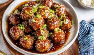 A bowl of sweet and sour meatballs over steamed white rice drizzled with sticky hoisin-honey glaze and garnished with sliced green onions