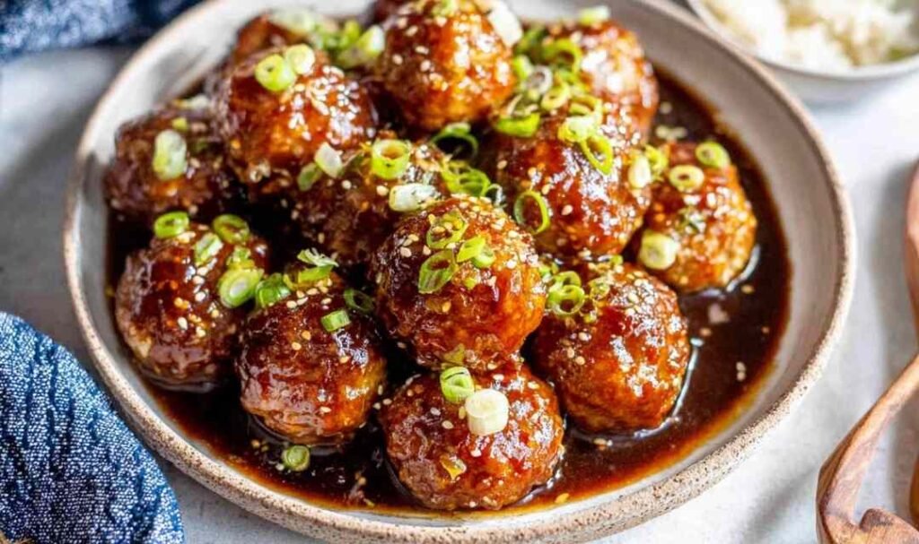A bowl of sweet and sour meatballs over steamed white rice drizzled with sticky hoisin-honey glaze and garnished with sliced green onions