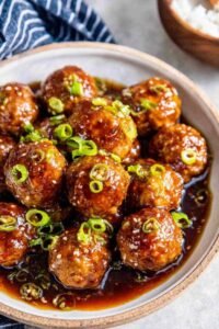 A bowl of sweet and sour meatballs over steamed white rice drizzled with sticky hoisin-honey glaze and garnished with sliced green onions