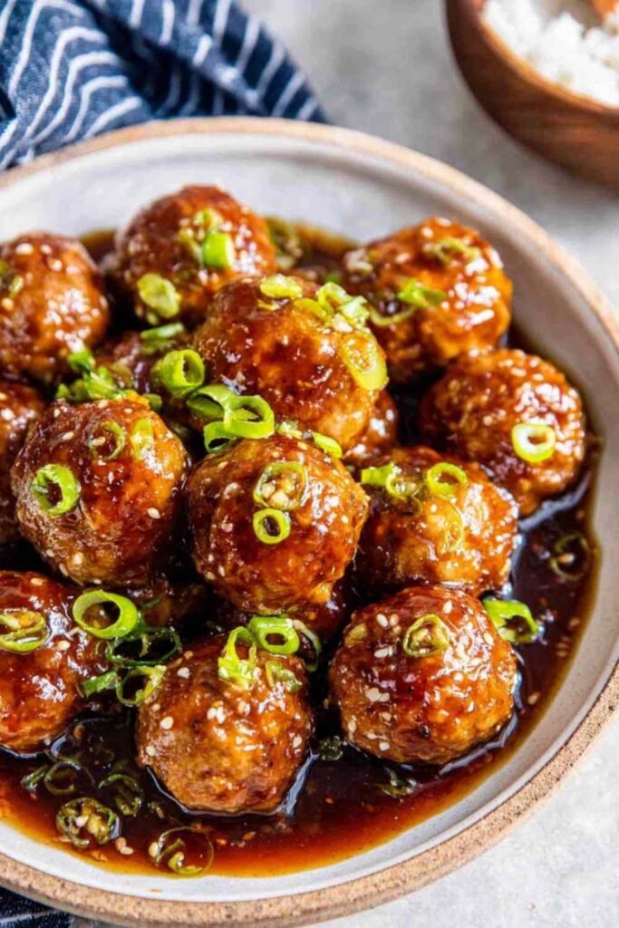 A bowl of sweet and sour meatballs over steamed white rice drizzled with sticky hoisin-honey glaze and garnished with sliced green onions