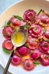 Valentine's Day ravioletti with pink beet pasta served in silky butter sauce with toasted walnuts and fresh mint and dill on a white plate