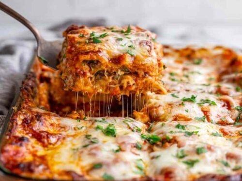 A slice of vegetable lasagna being lifted from a white baking dish showing distinct layers of creamy ricotta, marinara sauce, sauteed vegetables, and melted mozzarella cheese