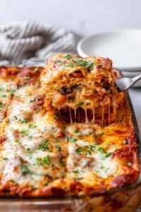 A slice of vegetable lasagna being lifted from a white baking dish showing distinct layers of creamy ricotta, marinara sauce, sauteed vegetables, and melted mozzarella cheese