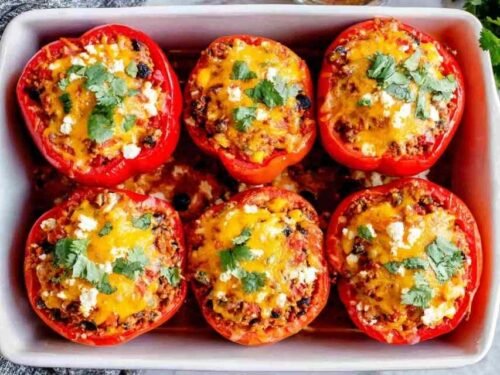 Six colorful vegetarian stuffed peppers in a baking dish with melted cheese and fresh cilantro garnish