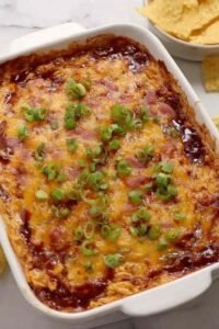 Baked BBQ chicken dip bubbling in a white baking dish topped with melted cheddar cheese and chopped green onions