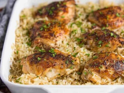 Baked chicken and rice casserole in a white 9x13 dish with golden seasoned chicken thighs over creamy rice, garnished with fresh parsley