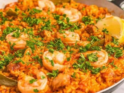 A large skillet of homemade chicken paella recipe with golden saffron rice, shrimp, chorizo, and fresh parsley on a wooden table