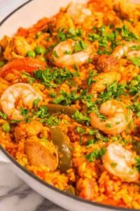 A large skillet of homemade chicken paella recipe with golden saffron rice, shrimp, chorizo, and fresh parsley on a wooden table